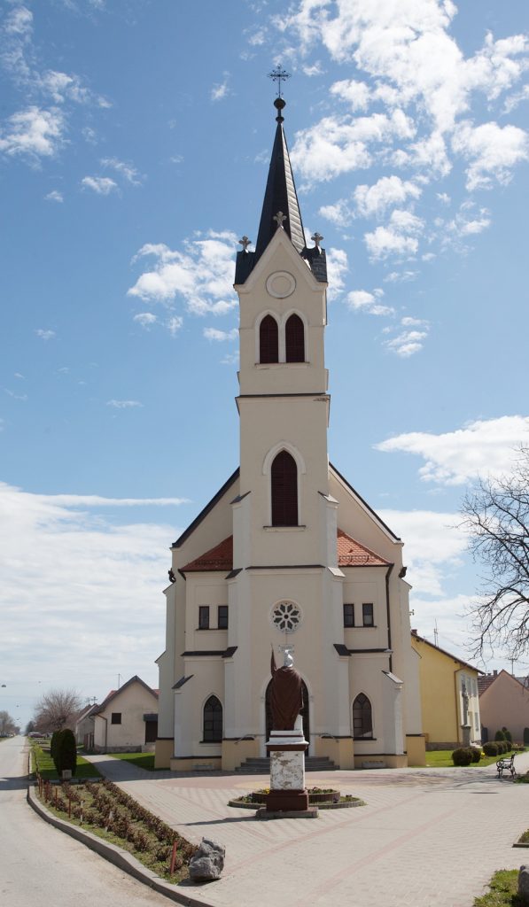 White light-beige church with a tall, pointed spire and arched windows, located in a paved plaza with a statue in front.