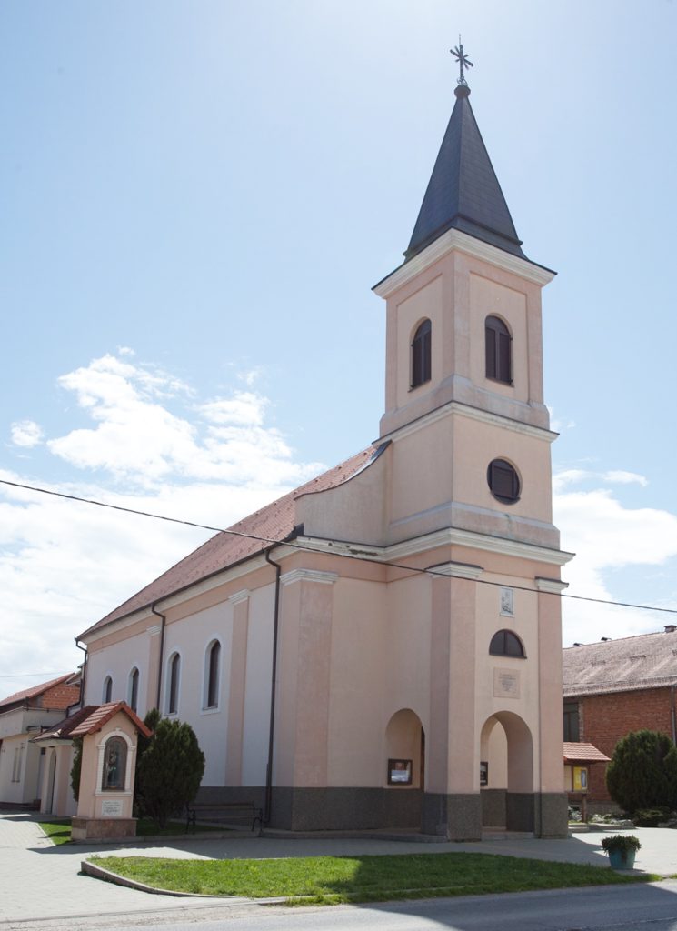 Pale pink church with a tall, dark steeple topped by a cross against a blue sky.