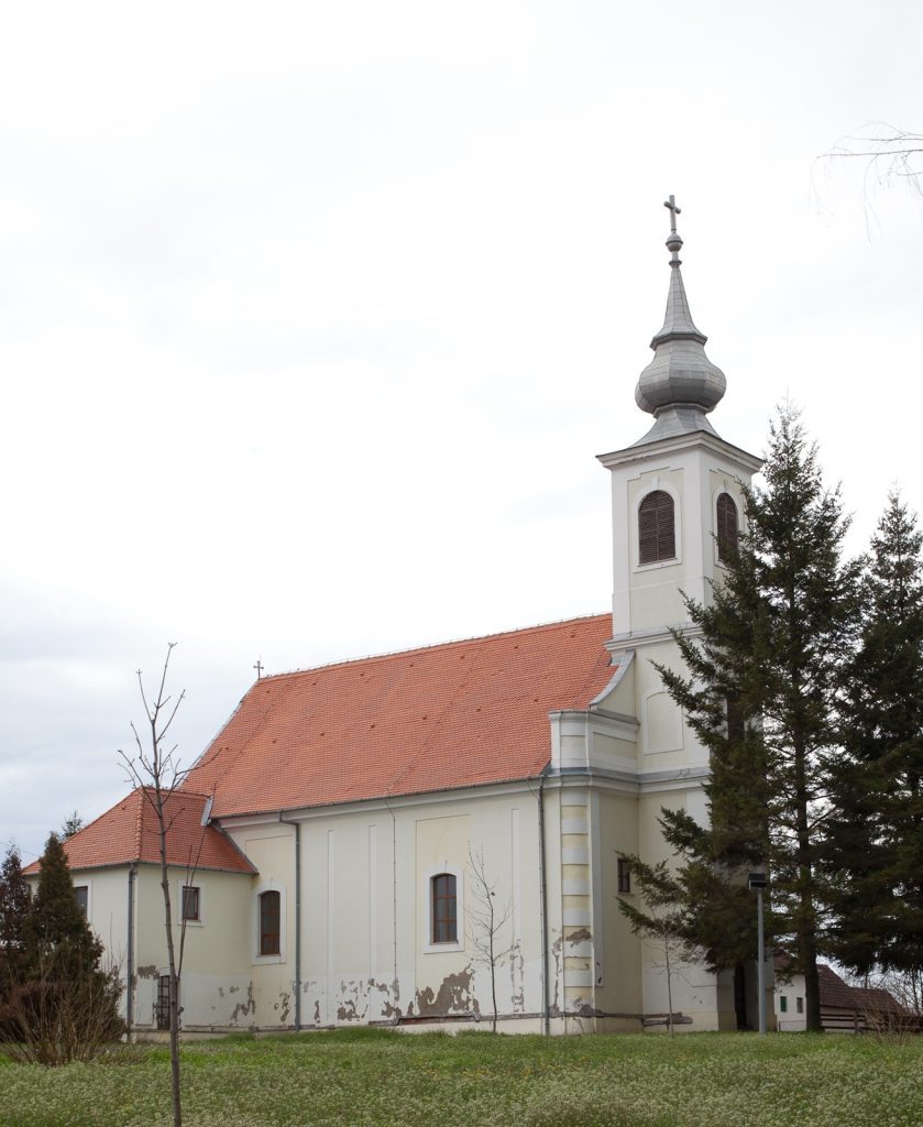 White church with a red-tiled roof and a tall steeple topped with a cross, flanked by evergreen trees.