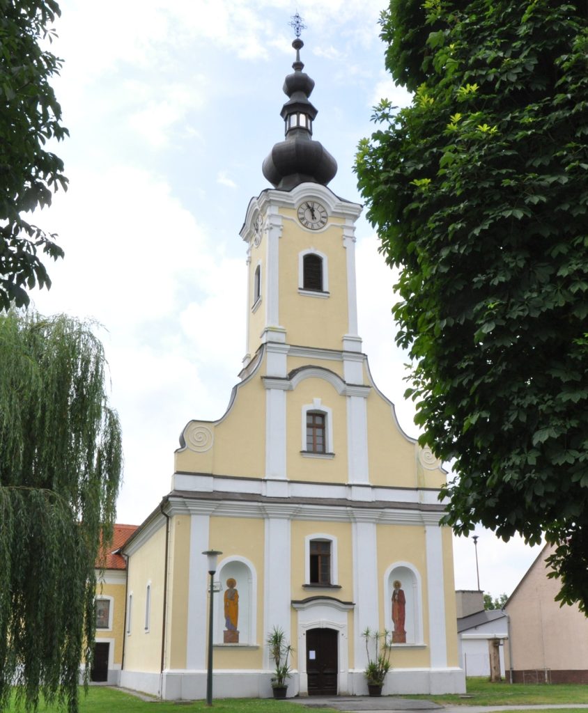 Front view of a yellow church with a clock tower, framed by green trees on both sides.