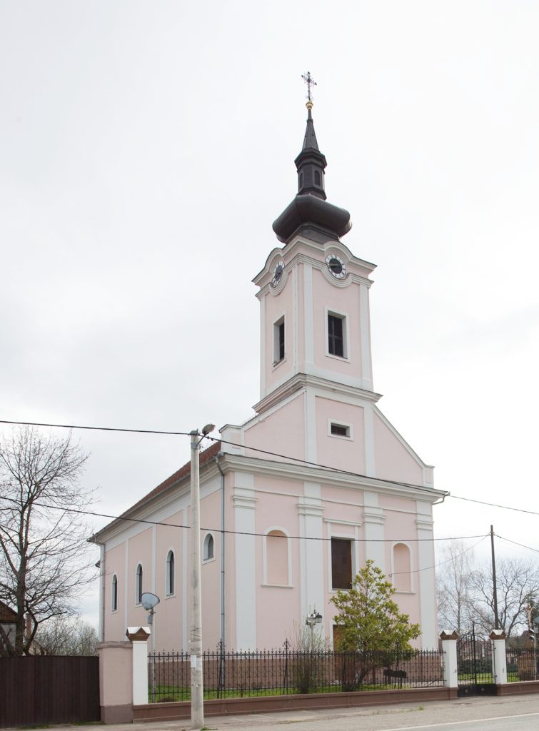Pink church with white trim and a tall black clock tower topped by a cross, beside a fence on a street.
