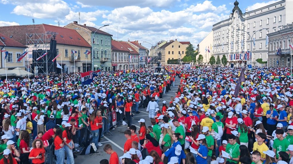 Large crowd of people in colorful shirts lines a city street for a parade, with flags and historic buildings nearby.