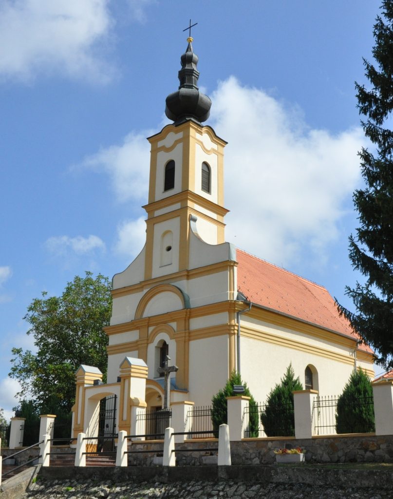 Church with a tall steeple and cross, cream and yellow facade, and a red-tiled roof set against a blue sky with trees nearby.