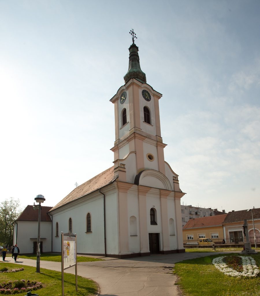 White church with a tall green steeple and clock faces reaching toward a clear blue sky; walkway and garden in the foreground.
