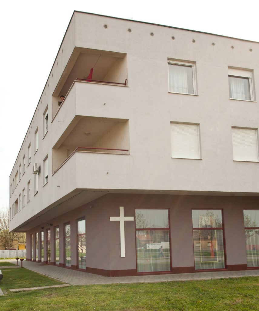 Modern three-story building with a large white cross on the dark ground-floor wall, glass storefronts, and balconies on the upper levels (one person on a balcony).