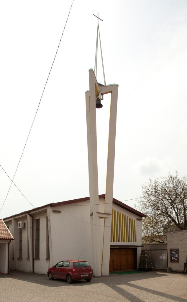 Modern bell tower rising beside a white building, with a cross at the top and a visible bell beneath it.