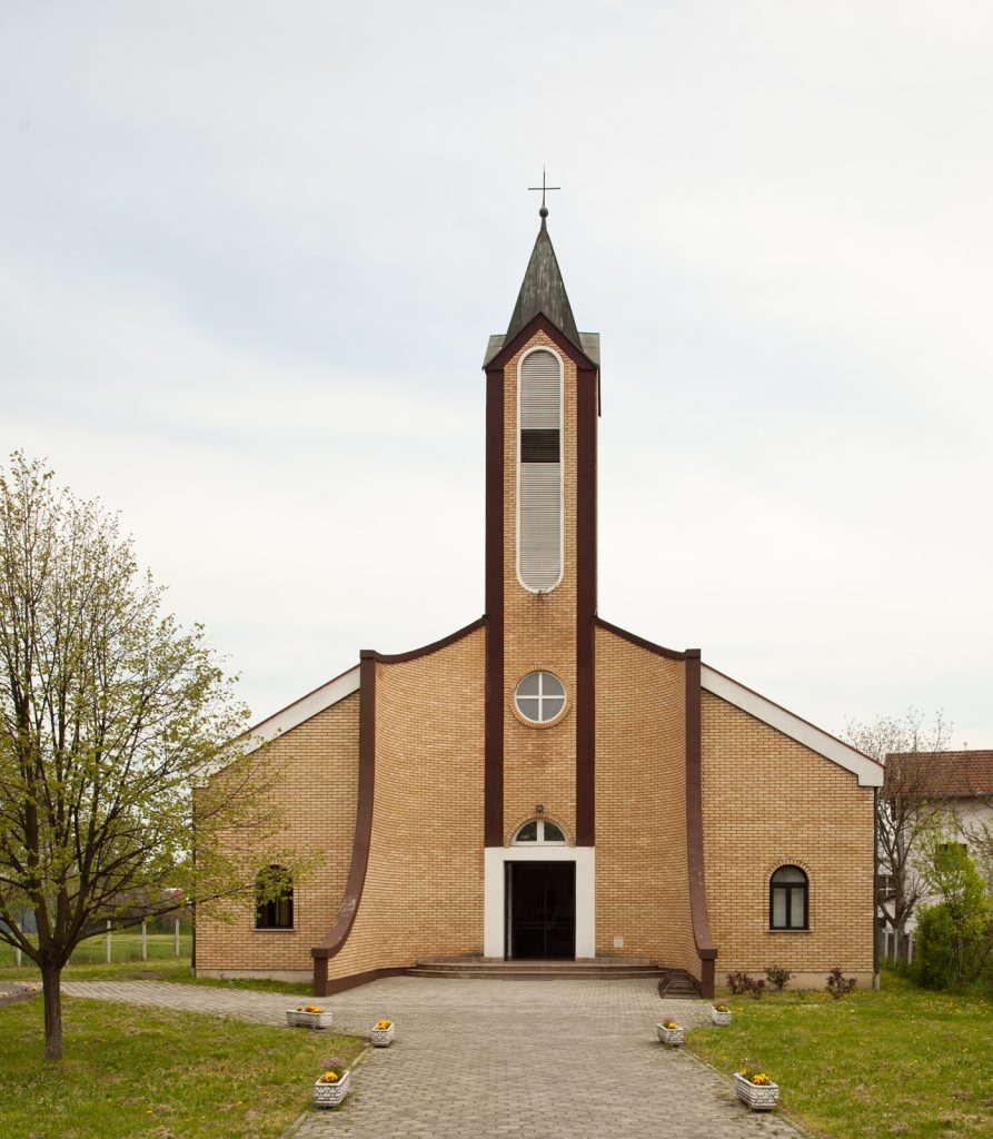 Brick church with tall central steeple and cross, arched door and windows, surrounded by a lawn and trees.