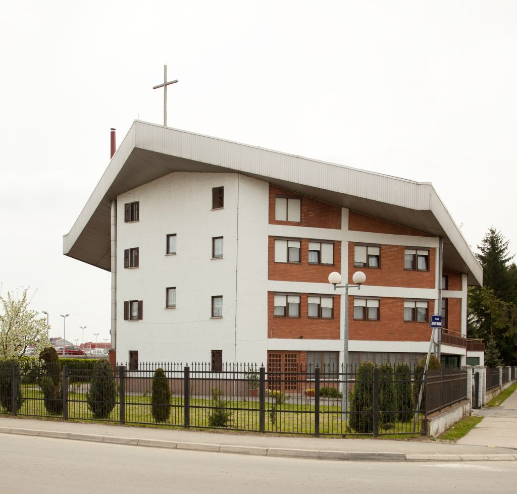 White and red-brick multi-story building with a sloped roof and a cross on top. The property is fenced and sits beside a street.