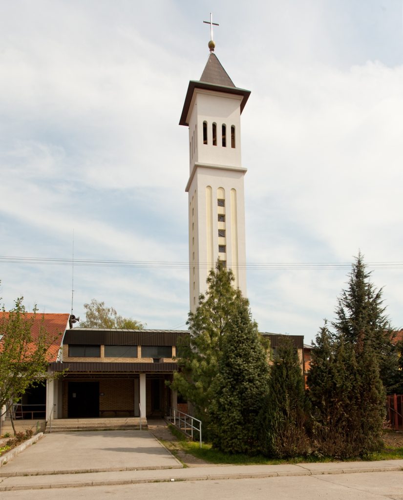 Tall white church bell tower with a cross on top, set against a blue sky, with a low building and evergreen trees nearby.