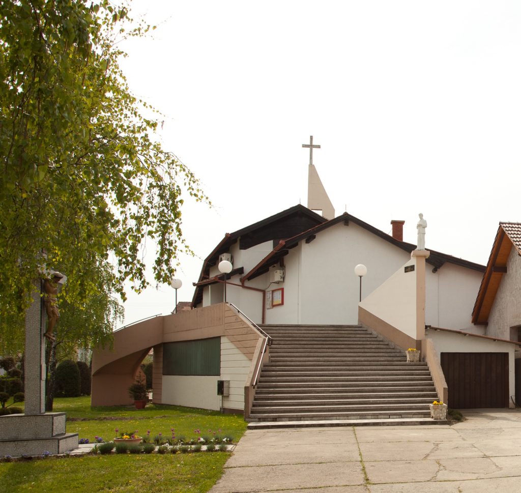 Front view of a white church with a cross on the steeple, wide concrete steps, and a statue on the right pedestal.