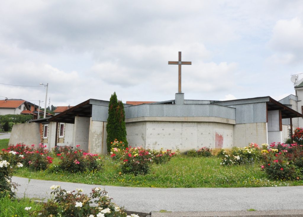Small concrete church with a wooden cross on top, metal siding, and a rose-filled foreground garden nearby.