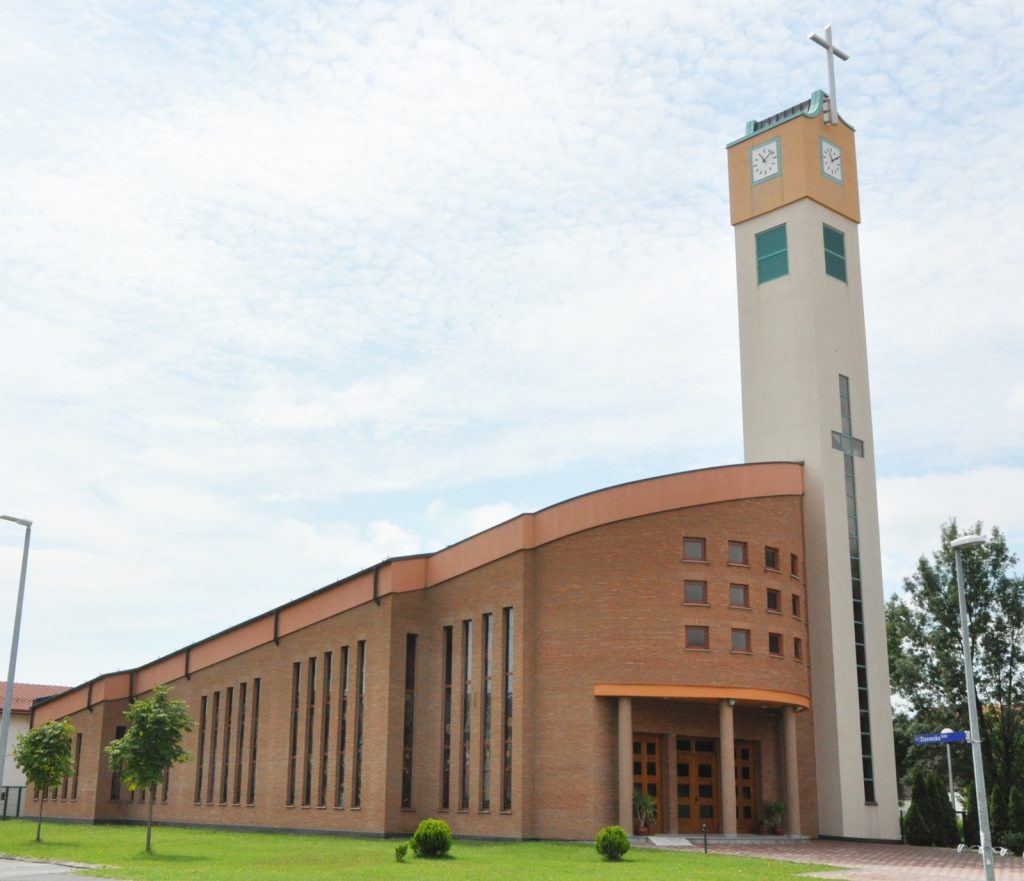 Front view of a brick church-like building with a tall beige clock tower topped by a cross against a blue sky.