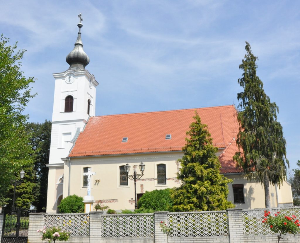 White church with a tall tower and onion dome, red-tiled roof, surrounded by trees under a blue sky.