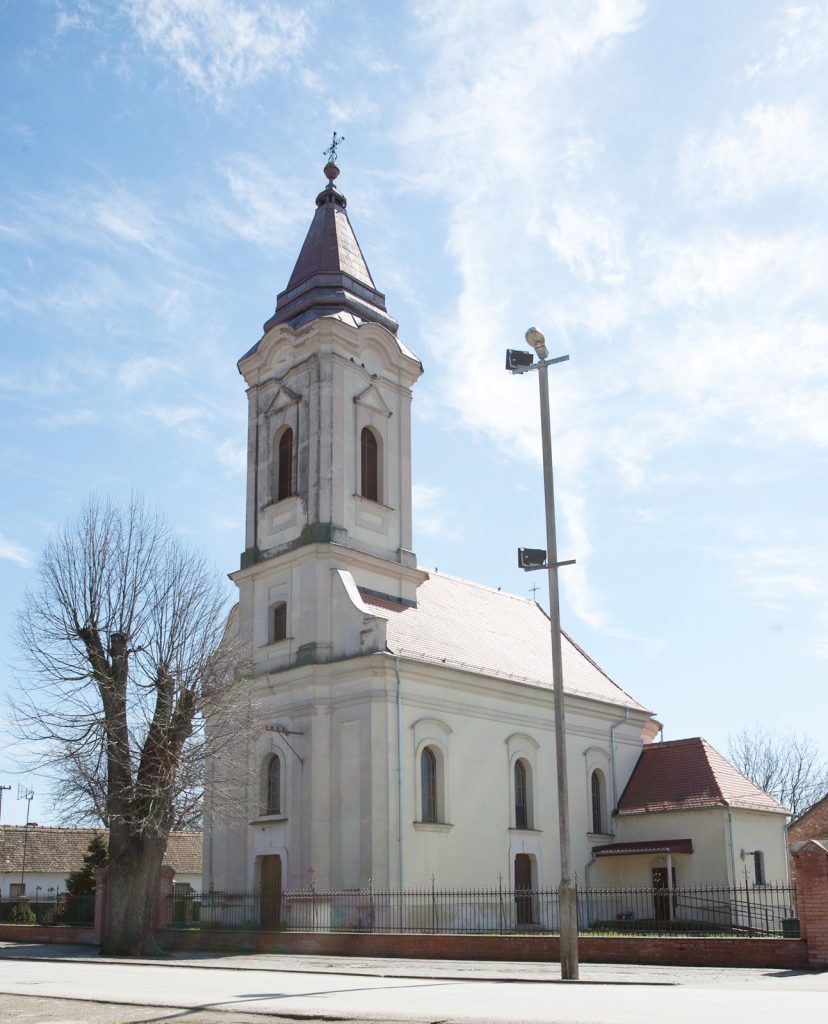 White church with a tall, pointed steeple against a bright blue sky; leafless trees and a street lamp in the foreground.