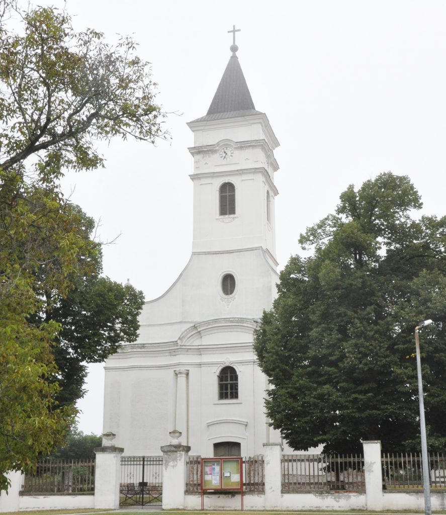 White church with a tall steeple topped by a cross, clock on tower, and surrounding trees behind a fence