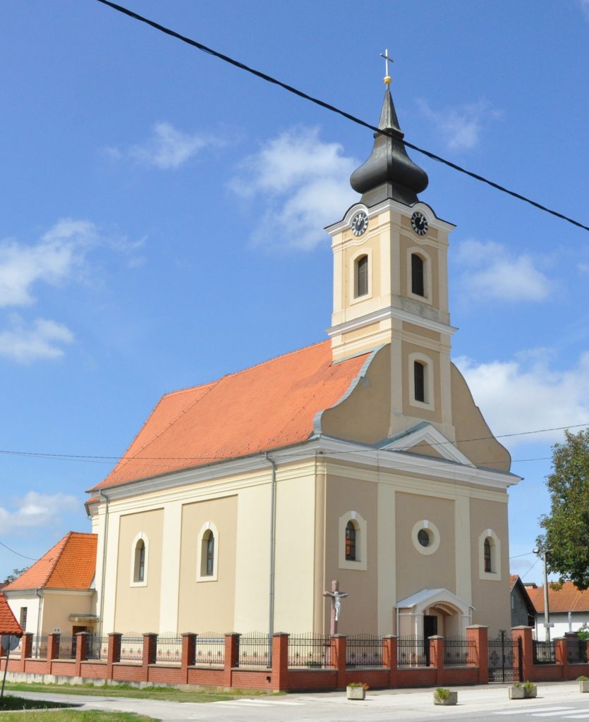 Church with beige walls, red-tiled roof, and a tall clock tower topped by a cross against a blue sky