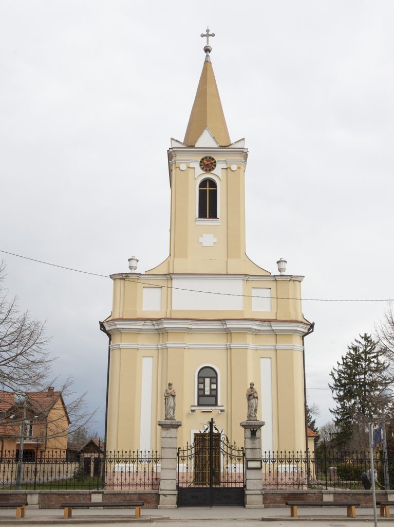 Yellow church with a tall steeple and cross, ornate gate at the entrance, and statues nearby.