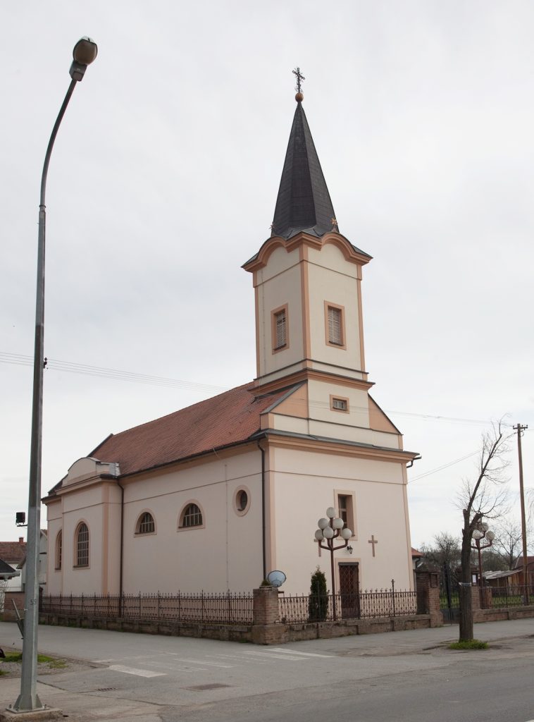 Church building with a tall dark spire and cross, beige facade, red roof, and a surrounding fence under a cloudy sky.