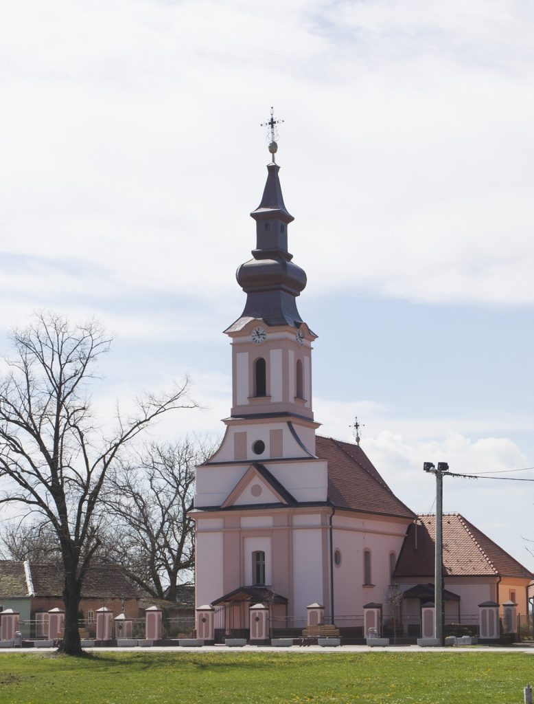 Pink church with a tall, multi-tiered spire and clock faces on the tower, set against a blue sky with leafless trees nearby