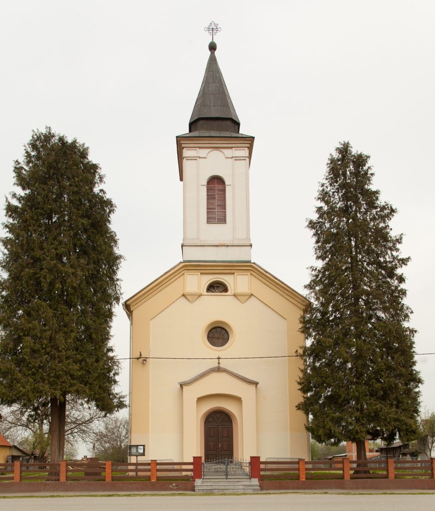 Church with tall steeple rising above its yellow facade, flanked by two evergreen trees and a low brick fence at the front.