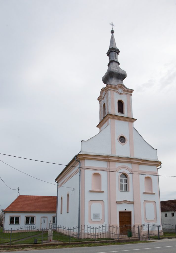 Pink and white church with a tall, ornate spire reaching into a cloudy sky in a small town setting.