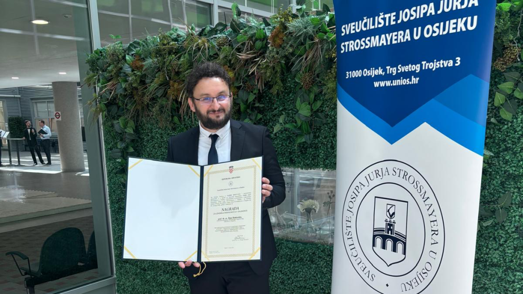 Man in a suit holding an open certificate folder, standing next to a blue university banner against a green plant wall.