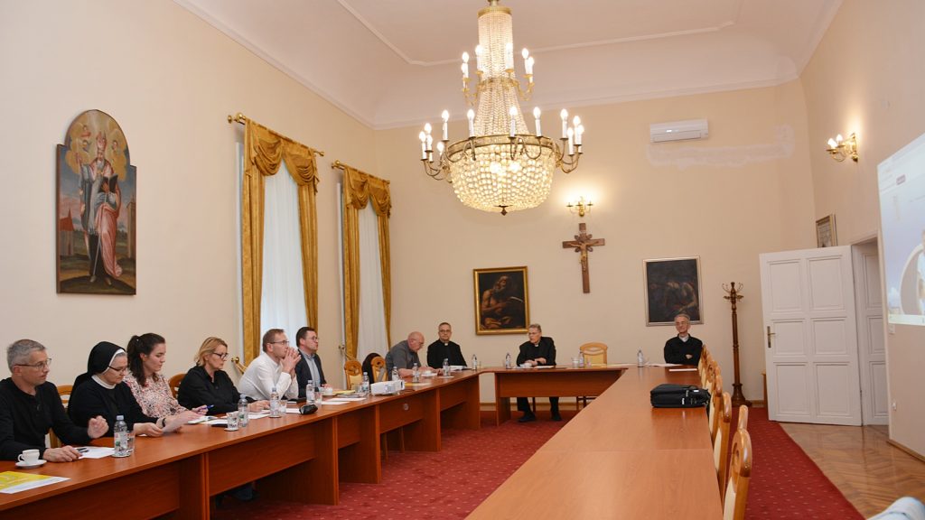 Meeting room in a church with clergy and participants seated around a long wooden table beneath a crystal chandelier.
