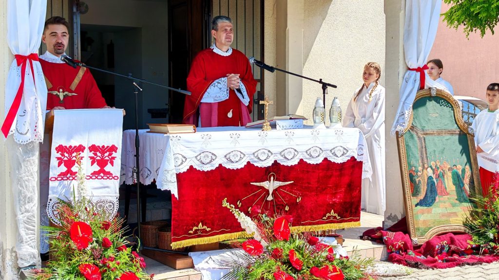 Outdoor Catholic altar scene with two priests in red vestments, white lace cuffs, at a lectern and altar covered in red-and-white cloths, surrounded by flowers.