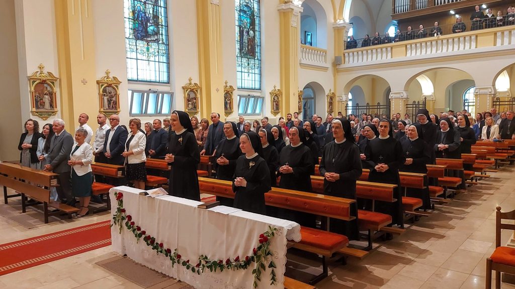 Church interior with nuns and parishioners standing at pews during a service; altar in foreground with white cloth and red flowersCreates a ceremonial mood.