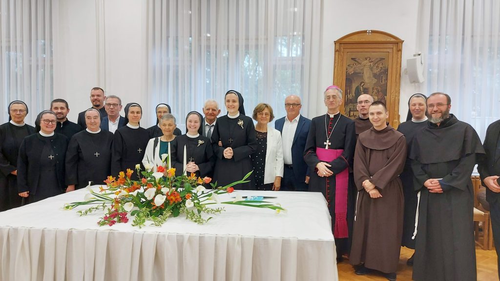 Group photo of Catholic nuns and priests gathered around a table with a floral centerpiece in a bright room with sheer curtains.