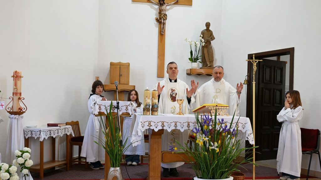 Catholic priests celebrate Mass at an altar with two children in white robes assisting, candles, and flowers in a bright church interior.