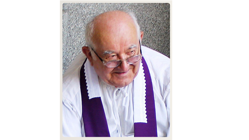 Older priest wearing a white shirt and purple stole, looking down and smiling.