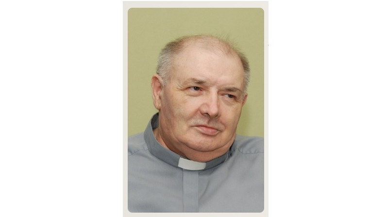 Portrait of an older man in a gray clerical shirt with a white clerical collar, looking to the side.