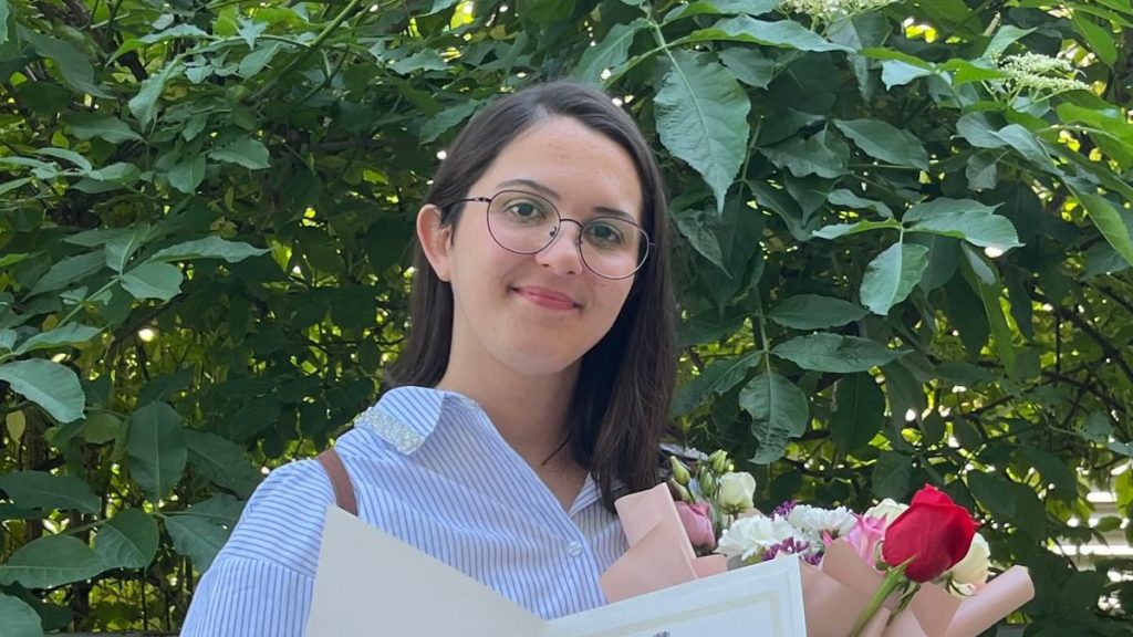 Smiling woman with round glasses holding a bouquet of flowers and a certificate outdoors in front of green foliage.