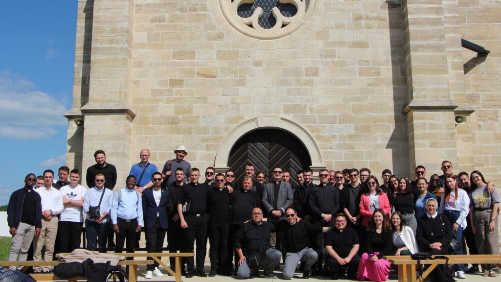 Large group portrait of adults standing in front of a stone church entrance with an arched door and circular window above.