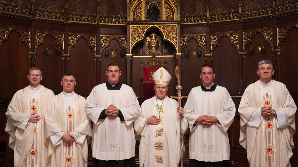 Group of six clergymen in white liturgical vestments posing in a richly carved church sanctuary, central bishop with staff wearing a mitre.