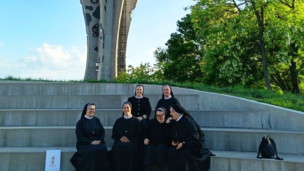 Six nuns in black habits sit on concrete steps outdoors near a tall monument, with green trees to the right and a backpack on the bench nearby.