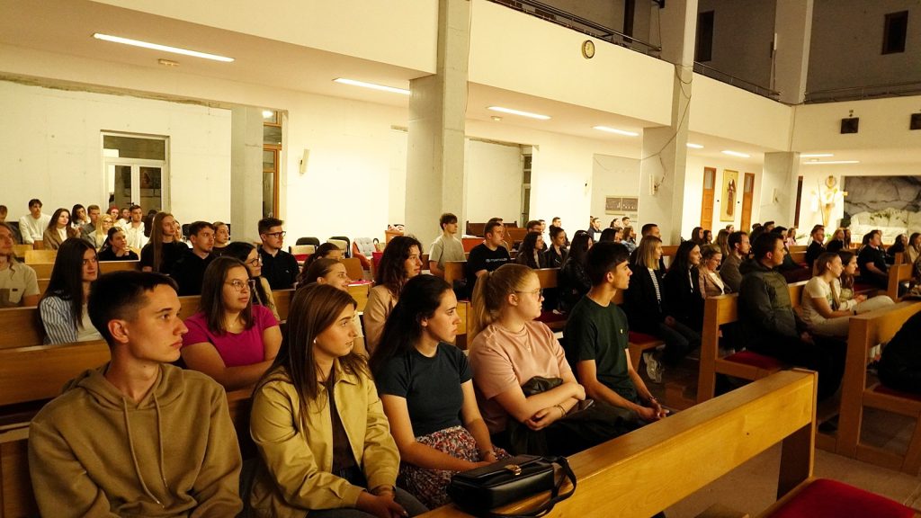 Audience of young adults seated in wooden pews inside a well-lit lecture hall during a presentation and listening attentively to a speaker. (informative)