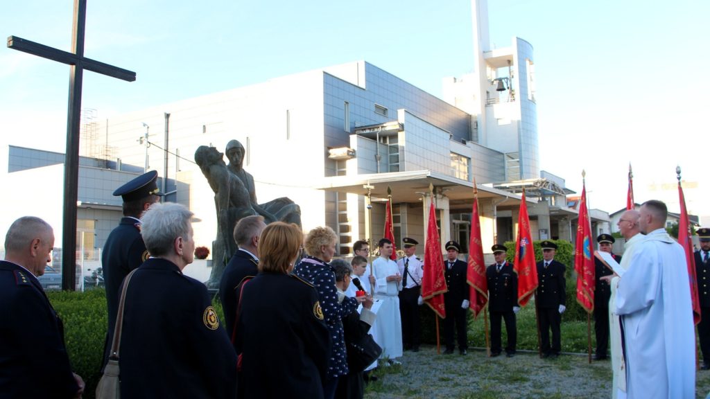 Group of uniformed veterans and civilians at an outdoor ceremony beside a metal statue, with red flags and a priest in white robes.
