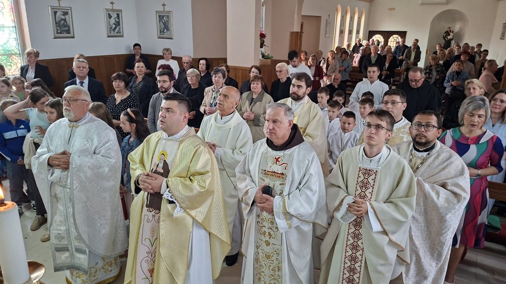 Priests in ornate white and gold vestments lead a church service at the front while a large congregation stands in pews behind them.