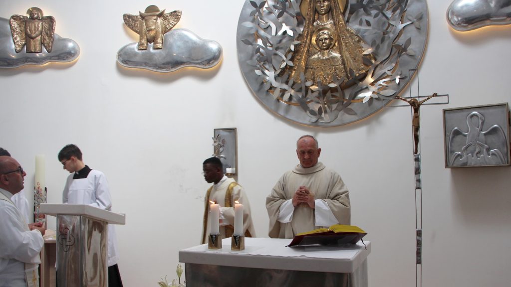 Priest in cream robes with hands folded at the altar during mass; candles lit and an open book nearby, with silver wall art behind.