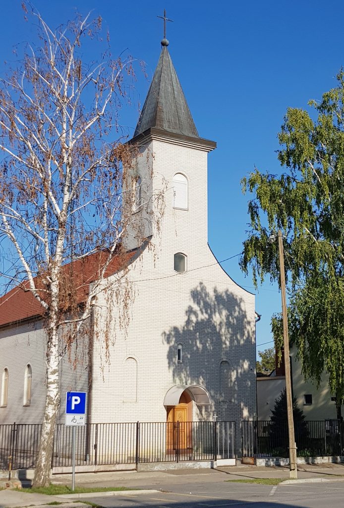 White brick church with a tall gray steeple topped by a cross, arched windows, and a wooden arched door, set against a clear blue sky with trees nearby and a fence in the foreground.