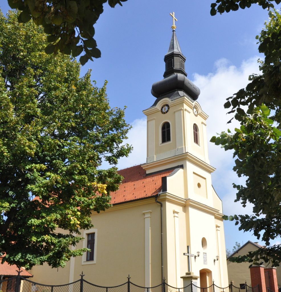 Church with a yellow facade and tall steeple framed by green trees against a blue sky.