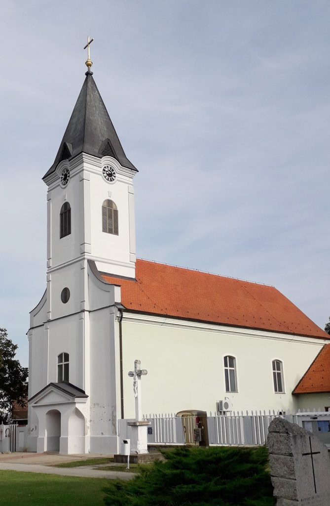White church with tall steeple featuring a cross atop, set against a blue sky and red-tiled roof