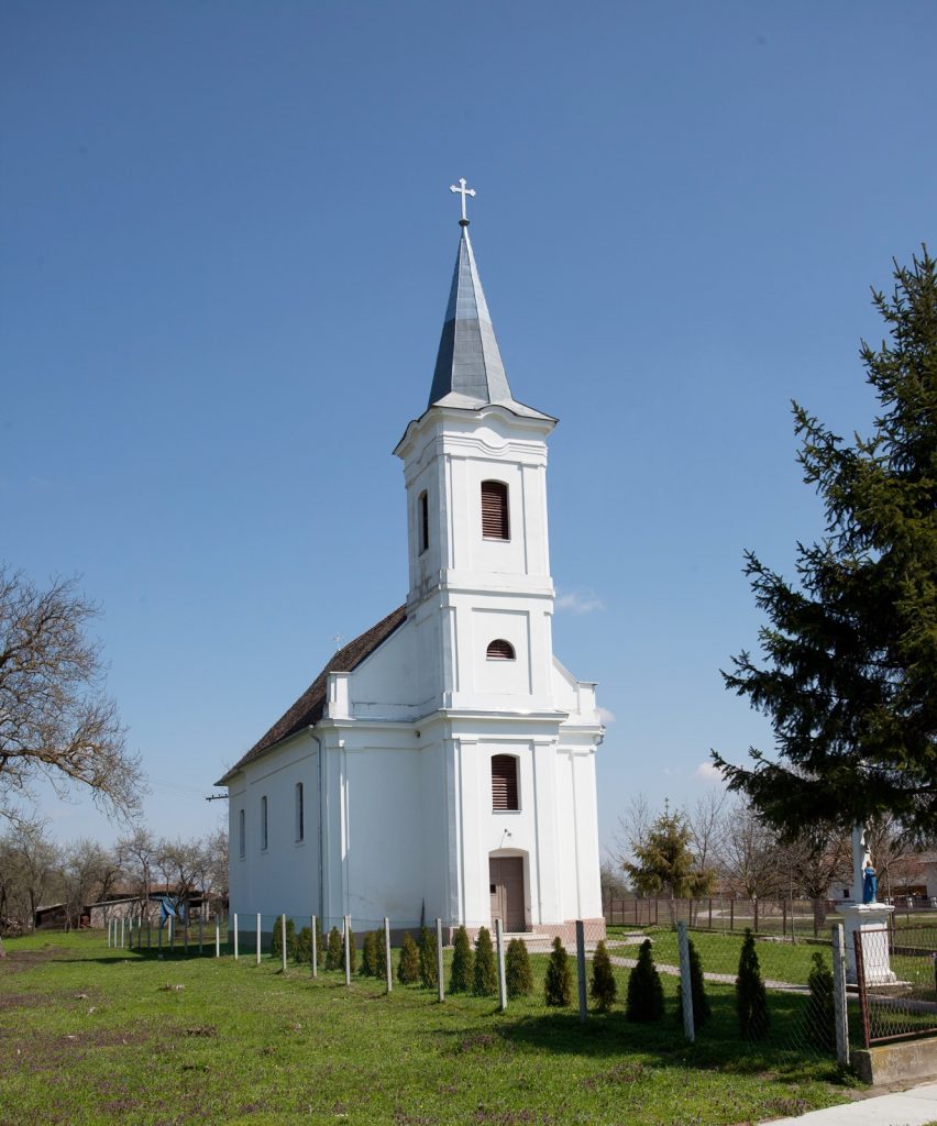 White church with a tall steeple and cross against a clear blue sky, enclosed by a fence with trees nearby.