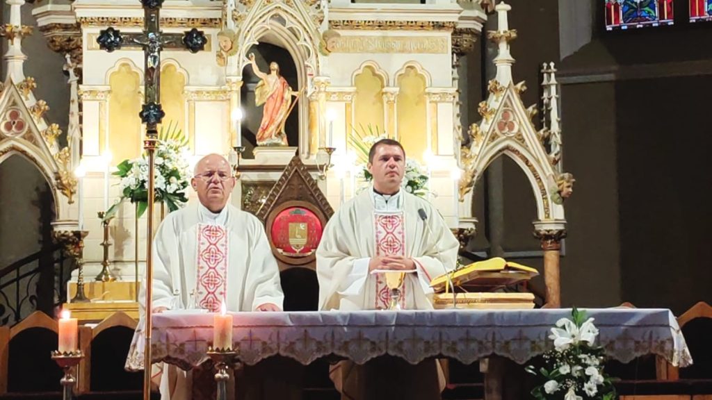 Two priests in white vestments stand at an ornate church altar during a service, with candles and floral arrangements behind them.