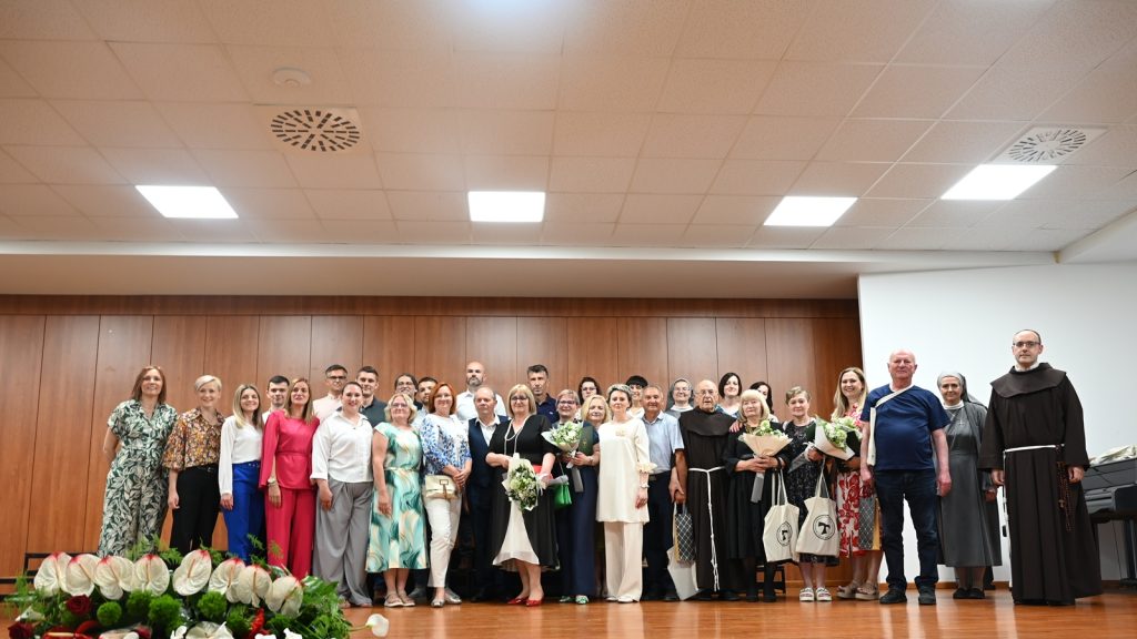 Large group portrait on a stage after a ceremony; attendees in formal attire, some holding bouquets, with monks on the right.