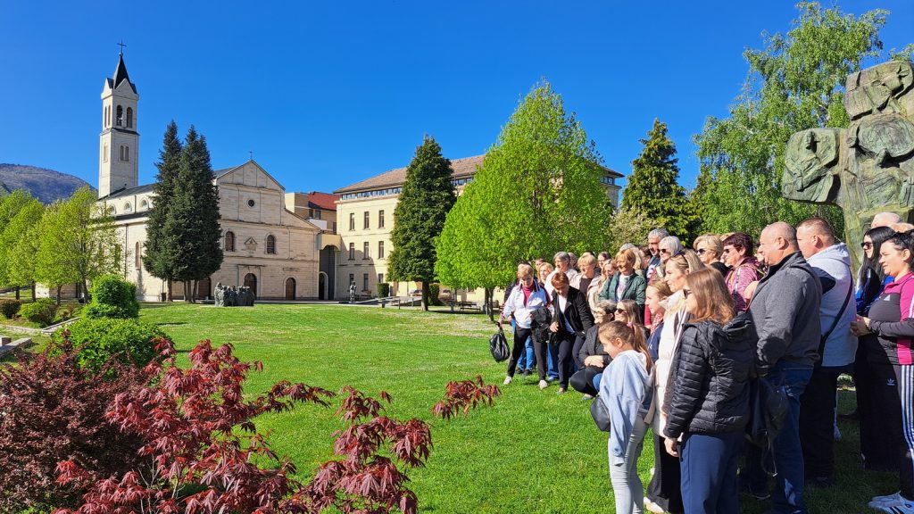 Group of adults and children standing on a green lawn outside a beige stone church with a tall bell tower under a bright blue sky.