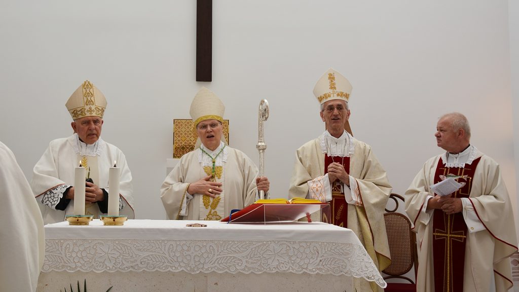 Four Catholic bishops in white and gold vestments stand at an altar during Mass, candles lit in front of them.