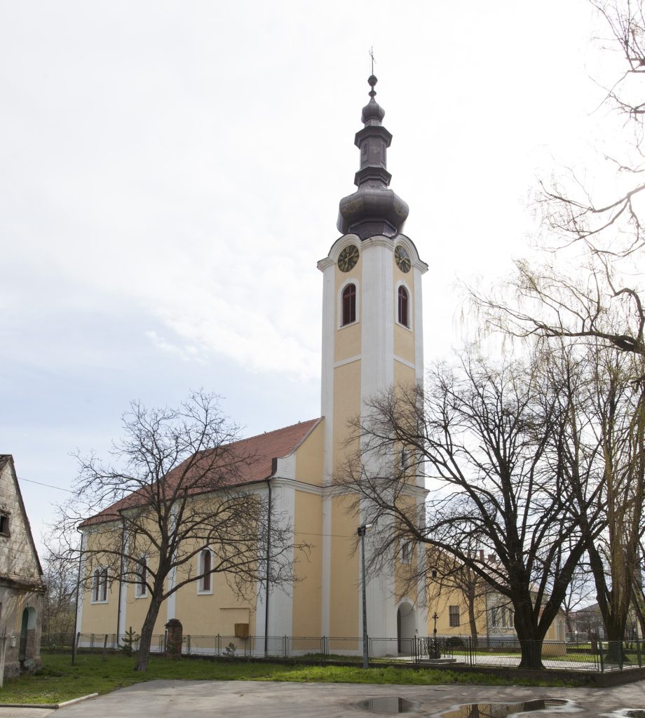 Tall church tower with a yellow facade and ornate, multi-tiered steeple, clock faces visible on the tower, surrounded by leafless trees.